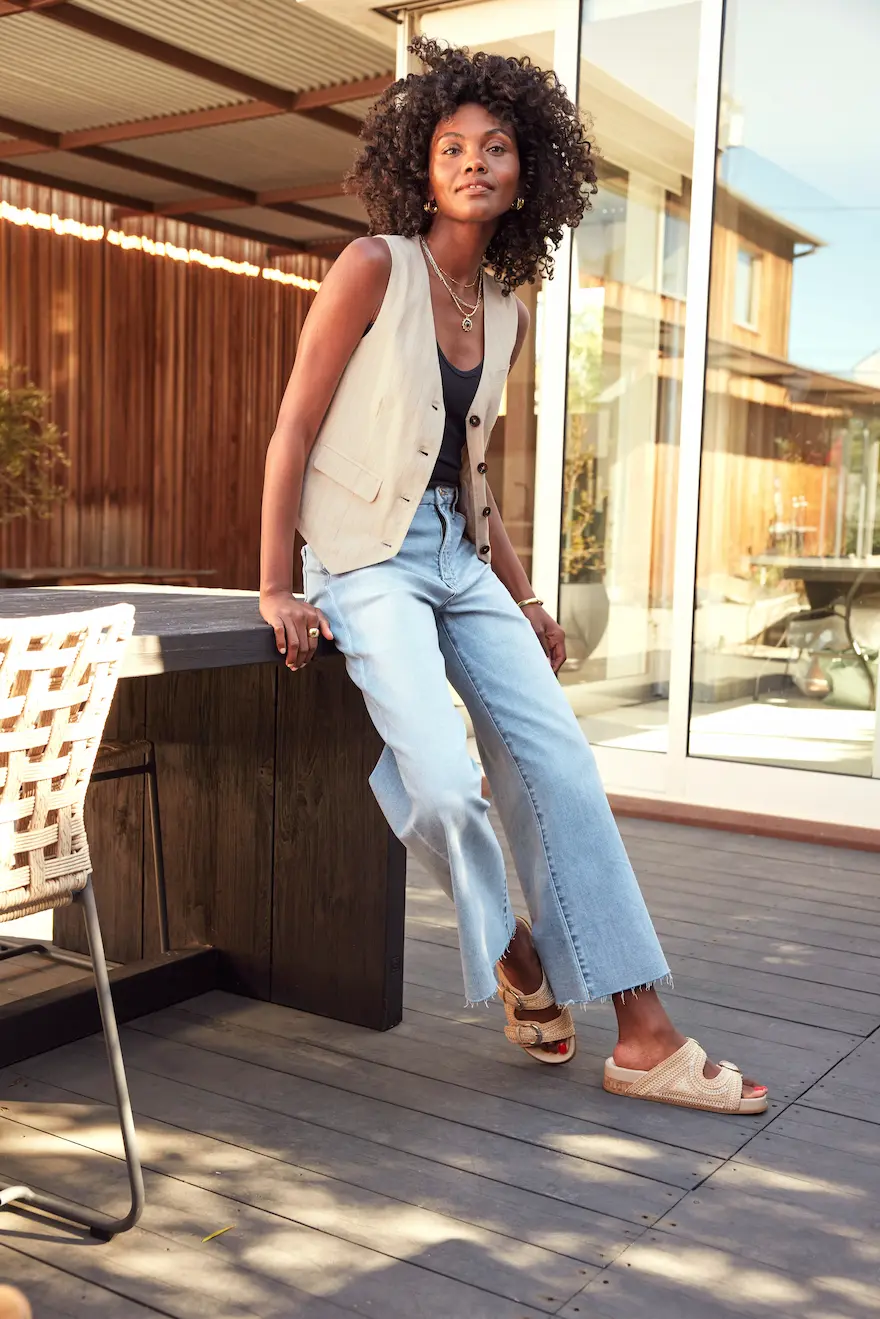 A woman with curly hair wearing an outfit consisting of a beige vest over a black top and light blue jeans leans against a wooden table in an outdoor setting. She is accessorized with gold jewelry and is wearing beige sandals. This look features a wide leg jeans outfit perfect for a relaxed outdoor environment.